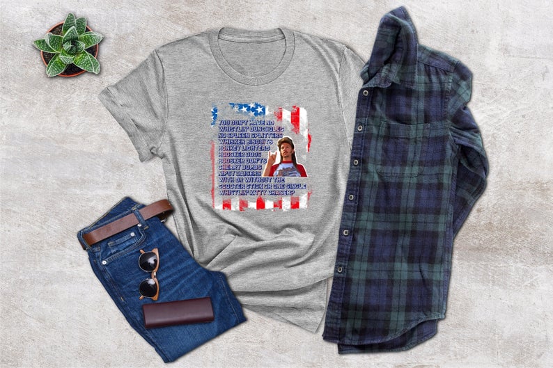 Op de afbeelding: Een grijs t-shirt met een afbeelding van een Amerikaanse vlag en tekst. De afbeelding bevat een spijkerbroek, een bruine riem, een zonnebril, een portemonnee en een geruit overhemd. Een kleine potplant is ook zichtbaar.