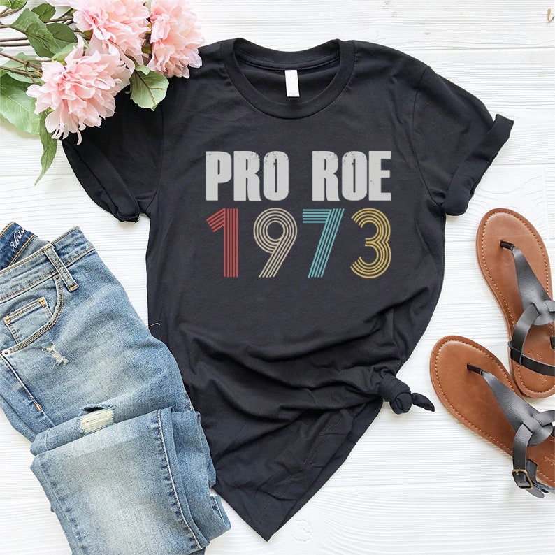 May include: Black t-shirt with the text "PRO ROE 1973" in a vintage style font with colorful stripes. The numbers are in red, blue, yellow, and green.