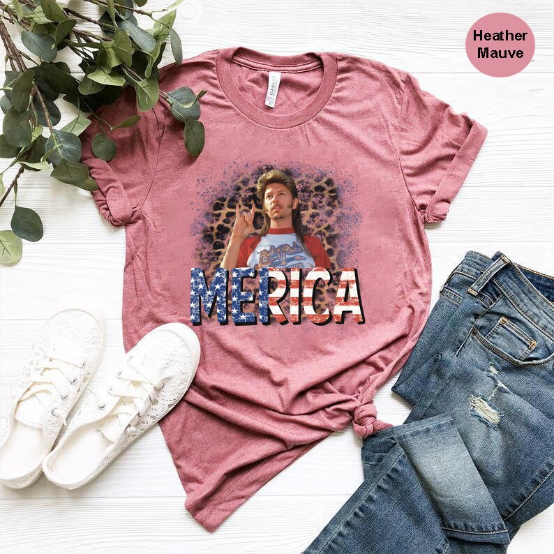 May include: A heather mauve t-shirt with a graphic of a man with a mullet and a beard wearing a red shirt with the word "MERICA" printed in red, white, and blue.