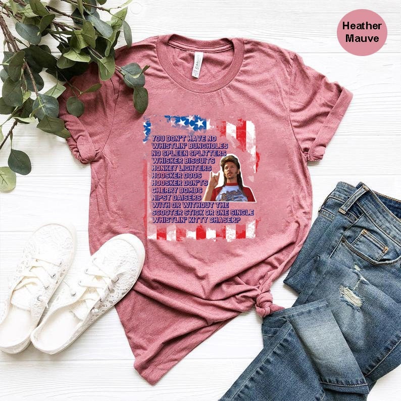 Op de afbeelding: Heather mauve t-shirt met een afbeelding met een man en tekst in een patriottisch ontwerp. De tekst luidt: "YOU DON'T HAVE NO WHISTLIN' BUNGHOLES...WHISTLIN' KITTY CHASER?" Het shirt wordt gecombineerd met een blauwe jeans en witte sneakers.