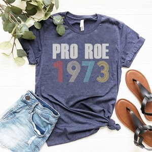 May include: A navy blue t-shirt with the text "PRO ROE 1973" in white with colorful retro-style numbers.