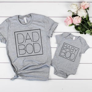 以下が含まれることがあります： 「DAD BOD」と「BABY BOD」の文字が黒い四角で囲まれた、大人用と乳児用の2枚のグレーのTシャツ。ピンクと白のバラの花束が白い木の表面に置かれています。