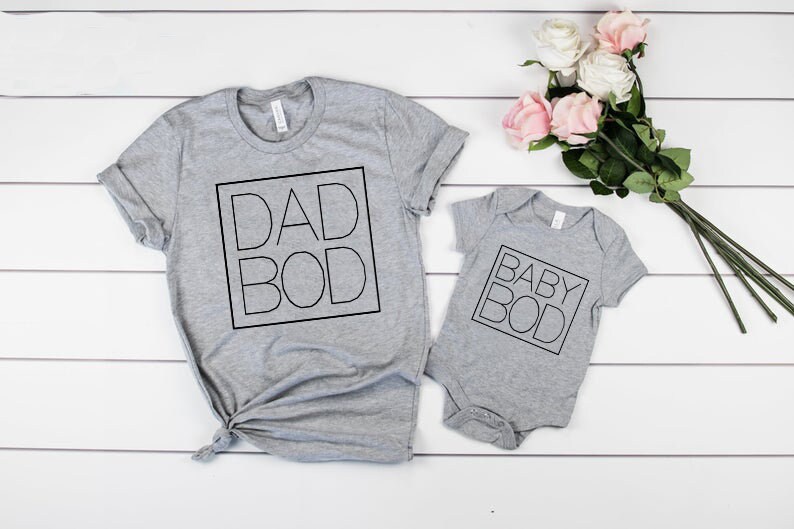 以下が含まれることがあります： 「DAD BOD」と「BABY BOD」の文字が黒い四角で囲まれた、大人用と乳児用の2枚のグレーのTシャツ。ピンクと白のバラの花束が白い木の表面に置かれています。