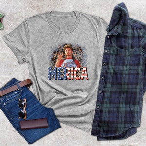 May include: A gray t-shirt with a graphic of a man with a mustache and a beard wearing a red, white, and blue shirt with the word "MERICA" printed in red, white, and blue letters.
