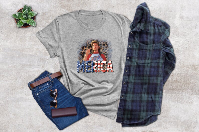 May include: A gray t-shirt with a graphic of a man with a mustache and a beard wearing a red, white, and blue shirt with the word "MERICA" printed in red, white, and blue letters.