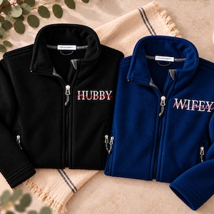 May include: A black fleece jacket with the word HUBBY printed in white and a blue fleece jacket with the word WIFEY printed in white. Both jackets feature full-length front zippers, high collars, and two zippered side pockets.