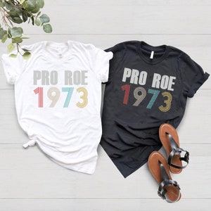 May include: Two t-shirts with the text "Pro Roe 1973" in a retro font. The shirts are white and black and the numbers are in red, blue, and yellow.