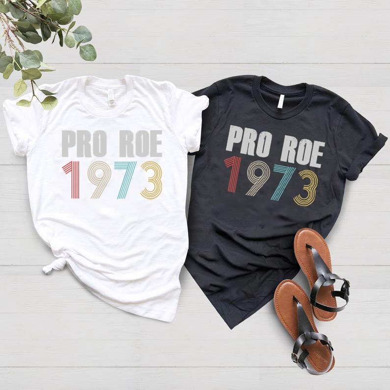 May include: Two t-shirts with the text "Pro Roe 1973" in a retro font. The shirts are white and black and the numbers are in red, blue, and yellow.