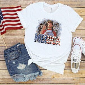 May include: White t-shirt with a red, white, and blue graphic of a man with a beard and the word "Merica" printed on it. The shirt is paired with denim shorts and a pair of white sneakers.