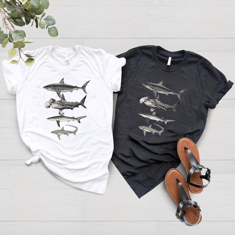 May include: Two t-shirts, one white and one black, with a graphic print of five different types of sharks. The sharks are arranged vertically, starting with a great white shark at the top and ending with a hammerhead shark at the bottom.