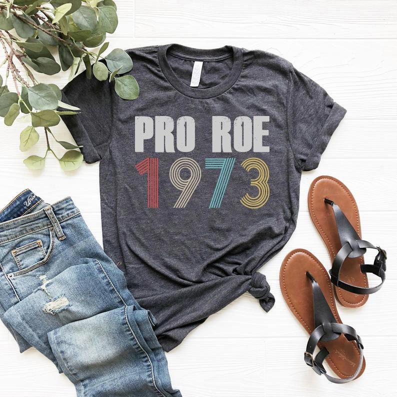 May include: A dark gray t-shirt with the text "PRO ROE 1973" in a vintage style font. The numbers are in different colors, with red, yellow, blue, and green.