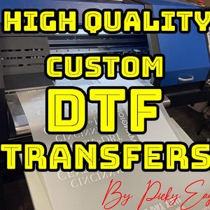 May include: A large blue and black printer with a white sheet of paper feeding through it. The paper has the text "HIGH QUALITY CUSTOM DTF TRANSFERS By Picky Eagle" printed on it in yellow letters.