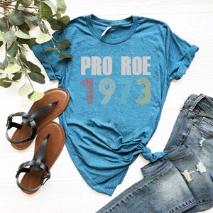 May include: A teal blue t-shirt with the text "PRO ROE 1973" in white and rainbow colored font. The shirt is laid out on a white surface with a pair of brown sandals and blue jeans.