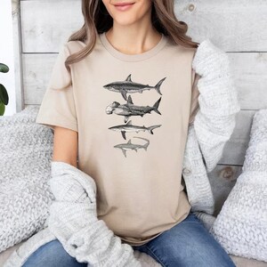 May include: A beige t-shirt with a graphic print of four different types of sharks. The sharks are in black and white and are arranged vertically on the shirt.