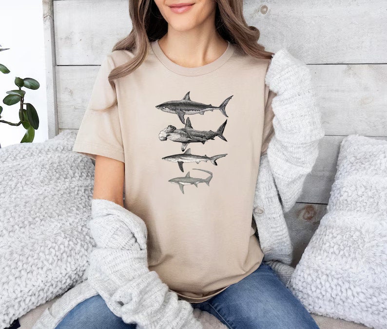May include: A beige t-shirt with a graphic print of four different types of sharks. The sharks are in black and white and are arranged vertically on the shirt.