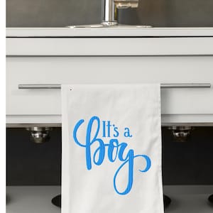 May include: White kitchen towel with the blue text "It's a Boy" embroidered on it. The towel is hanging from a silver bar in front of a white cabinet and a stainless steel faucet.