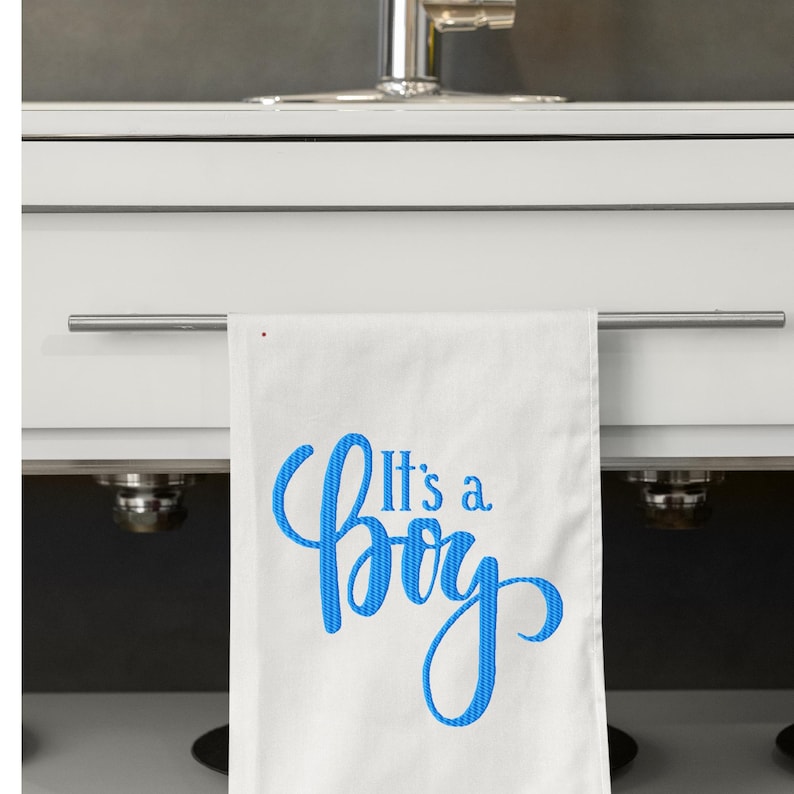 May include: White kitchen towel with the blue text "It's a Boy" embroidered on it. The towel is hanging from a silver bar in front of a white cabinet and a stainless steel faucet.