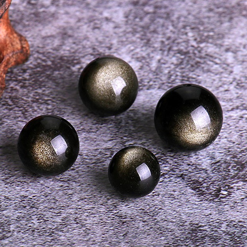 K&ouml;nnte beinhalten: Vier schwarze Obsidian-Kugeln mit goldenen Sprenkeln, angeordnet auf einer strukturierten grauen Oberfl&auml;che.
