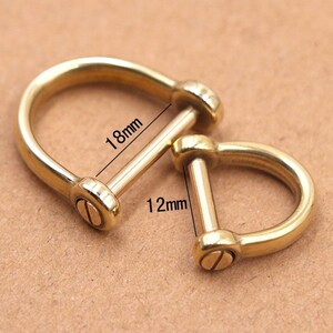 Op de afbeelding: Twee messing D-sluitingen. De ene meet 18 mm lang, de andere 12 mm. De sluitingen hebben een gebogen D-vorm met een verwijderbare pin die met een schroef is bevestigd. De achtergrond is lichtbruin.