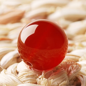 Könnte beinhalten: Eine leuchtend rote Glaskugel, die Licht reflektiert, liegt auf einer gewebten Oberfläche.