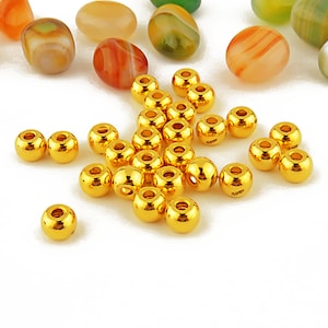 May include: A group of 25 gold-colored round beads, scattered on a white surface. The beads are shiny and have a smooth finish.