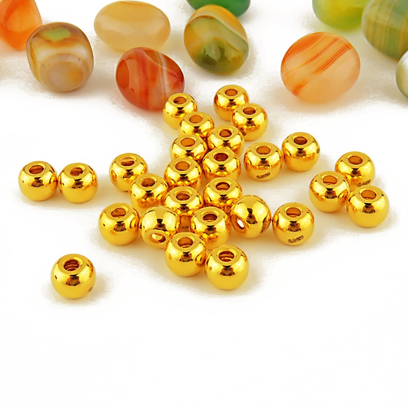 May include: A group of 25 gold-colored round beads, scattered on a white surface. The beads are shiny and have a smooth finish.
