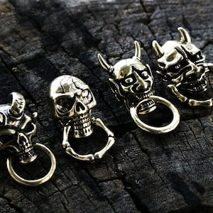Puede incluir: Cinco llaveros de latón con diferentes diseños de calaveras y demonios. Cada llavero tiene un anillo y un diseño tridimensional detallado.