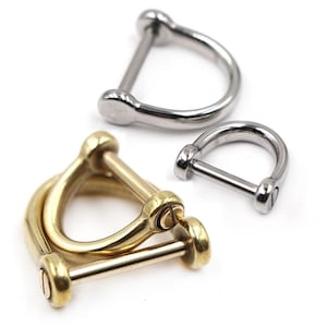 Op de afbeelding: Close-up van drie metalen D-sluitingen. Twee zijn zilverkleurig en &eacute;&eacute;n is goudkleurig. De D-sluitingen zijn gemaakt van metaal en hebben een gebogen D-vorm met een pin-sluiting. De goudkleurige sluiting is dubbel.
