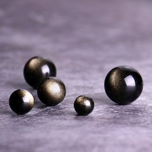 K&ouml;nnte beinhalten: F&uuml;nf schwarze Obsidian-Kugeln mit goldenen Sprenkeln, in einer Reihe auf einer grauen Oberfl&auml;che angeordnet. Die Kugeln sind in verschiedenen Gr&ouml;&szlig;en.