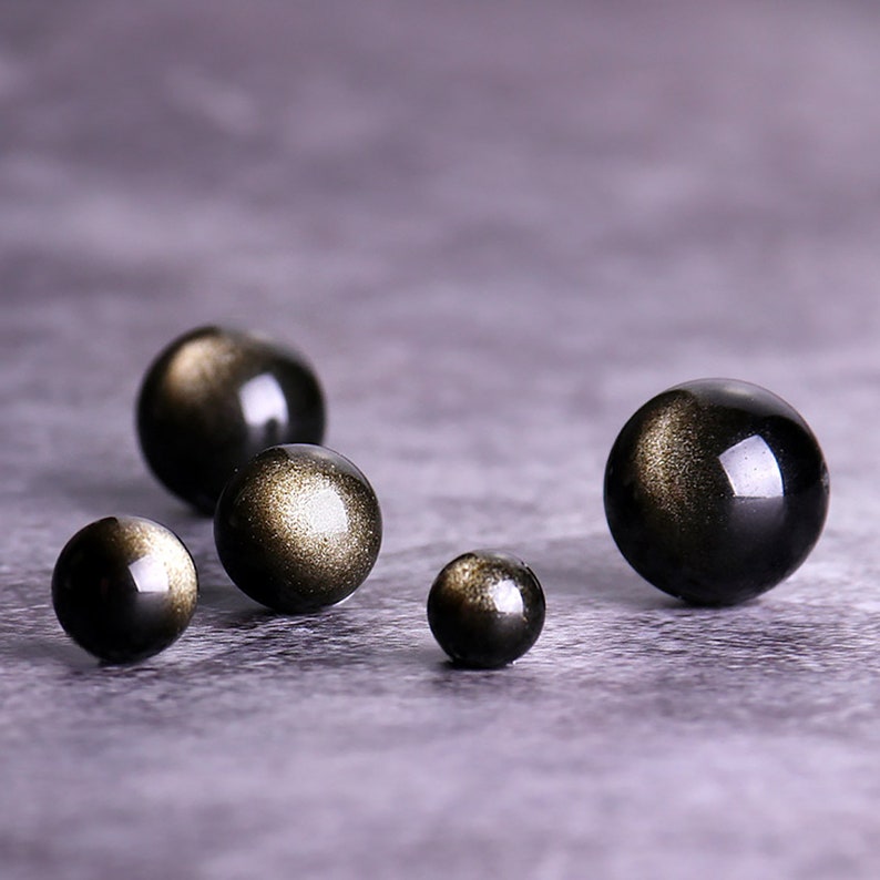 K&ouml;nnte beinhalten: F&uuml;nf schwarze Obsidian-Kugeln mit goldenen Sprenkeln, in einer Reihe auf einer grauen Oberfl&auml;che angeordnet. Die Kugeln sind in verschiedenen Gr&ouml;&szlig;en.