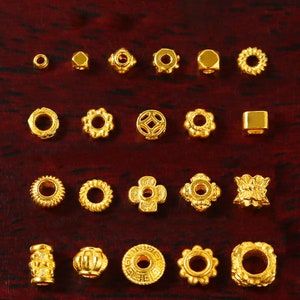 May include: A collection of 20 gold-colored metal beads with various shapes and designs, including round, square, and flower-shaped beads.