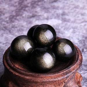 K&ouml;nnte beinhalten: F&uuml;nf schwarze Obsidian-Kugeln mit goldenen Sprenkeln, die in Pyramidenform auf einem Holzst&auml;nder angeordnet sind.