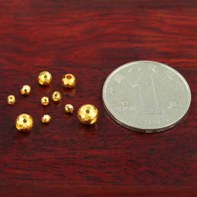 Op de afbeelding: Een verzameling gouden metalen kralen in verschillende maten, getoond op een houten oppervlak naast een Chinese yuan-munt.