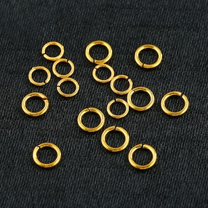 May include: A collection of 15 gold jump rings, scattered on a black fabric background. The jump rings are small and circular, perfect for jewelry making.