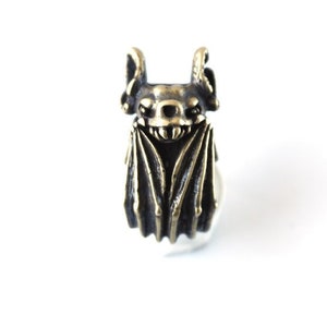 May include: A bronze bat ring with a detailed design. The bat has large ears and wings, and a toothy grin.