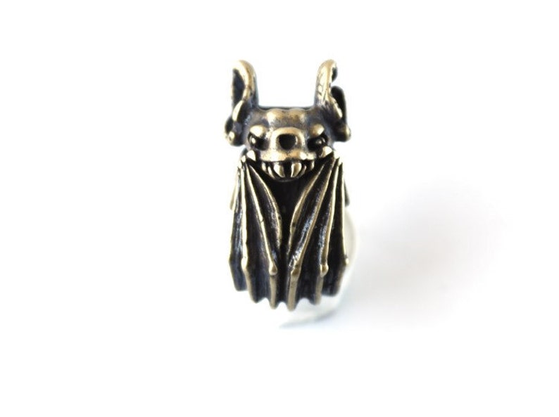 May include: A bronze bat ring with a detailed design. The bat has large ears and wings, and a toothy grin.