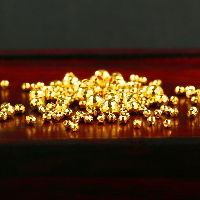 Op de afbeelding: Een stapel ronde gouden kralen met een getextureerd oppervlak. De kralen zijn verspreid over een donker rood houten oppervlak.