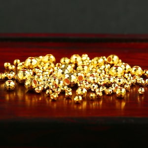 Op de afbeelding: Een stapel gouden metalen kralen met een getextureerd oppervlak. De kralen zijn rond en hebben een gat in het midden. De kralen liggen op een donker rood houten oppervlak.