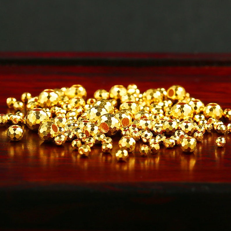 Op de afbeelding: Een stapel gouden metalen kralen met een getextureerd oppervlak. De kralen zijn rond en hebben een gat in het midden. De kralen liggen op een donker rood houten oppervlak.