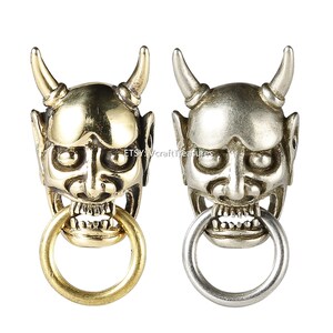 Könnte beinhalten: Zwei Schlüsselanhänger aus Metall mit einer detaillierten, stilisierten Dämonenmaske mit Hörnern und einem Ring im Maul. Ein Schlüsselanhänger ist golden, der andere ist silber.