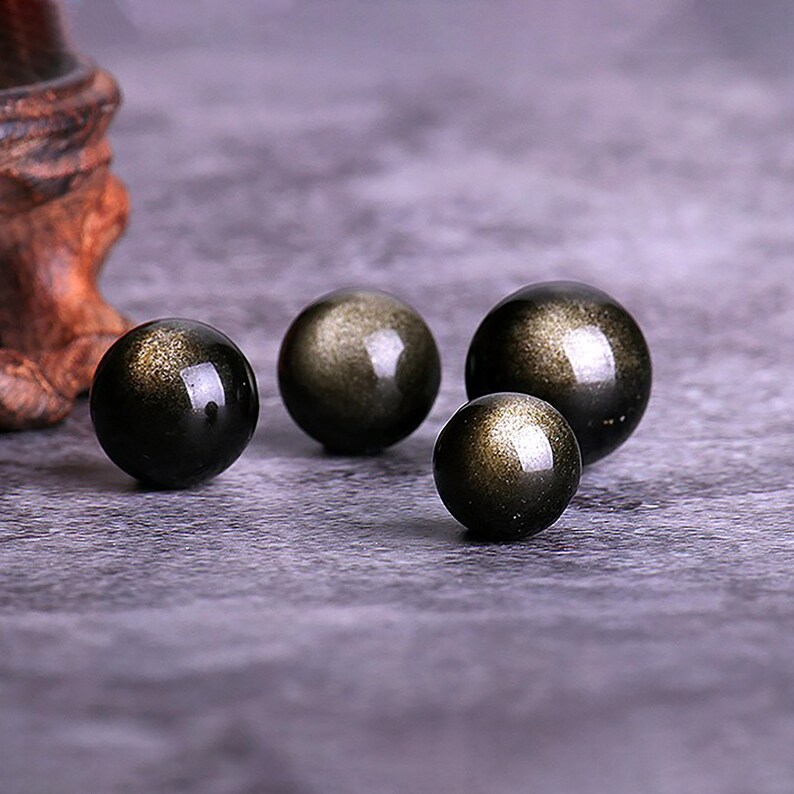 K&ouml;nnte beinhalten: Vier schwarze Obsidian-Kugeln mit goldenen Sprenkeln, angeordnet auf einer grauen Oberfl&auml;che. Die Kugeln sind unterschiedlich gro&szlig;.