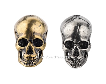 May include: Two metal skull beads, one gold and one silver, with detailed features. The skulls have black eye sockets and a toothy grin.