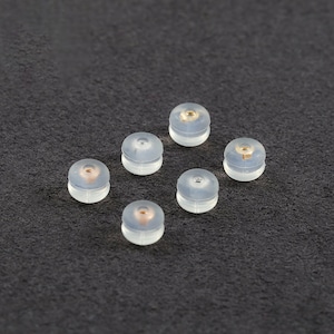 Peut inclure: Six dos d'oreille en plastique transparent avec un petit point doré au centre. Les dos sont disposés en demi-cercle sur une surface gris foncé.