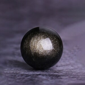 K&ouml;nnte beinhalten: Eine schwarze Obsidiankugel mit goldenen Flecken und einer reflektierenden Oberfl&auml;che.