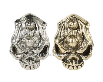 May include: Two skull-shaped metal beads, one silver and one gold, with three raised eyes on each bead.