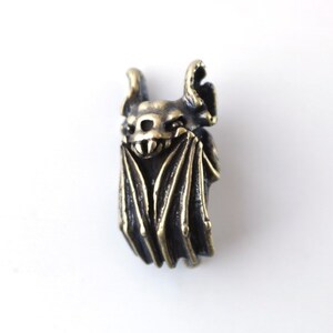 May include: A bronze bat charm with detailed wings and a menacing expression. The bat is hanging upside down with its wings spread out.