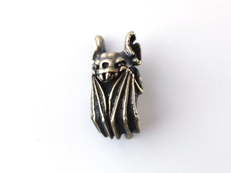 May include: A bronze bat charm with detailed wings and a menacing expression. The bat is hanging upside down with its wings spread out.