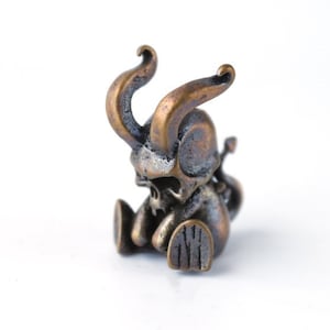 Puede incluir: Una pequeña figurita de bronce de un demonio sentado con cuernos y una cara de calavera.