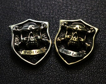 May include: Two silver and gold skull and crossbones pendants with a shield shape and the words "Good Luck" inscribed on a banner.