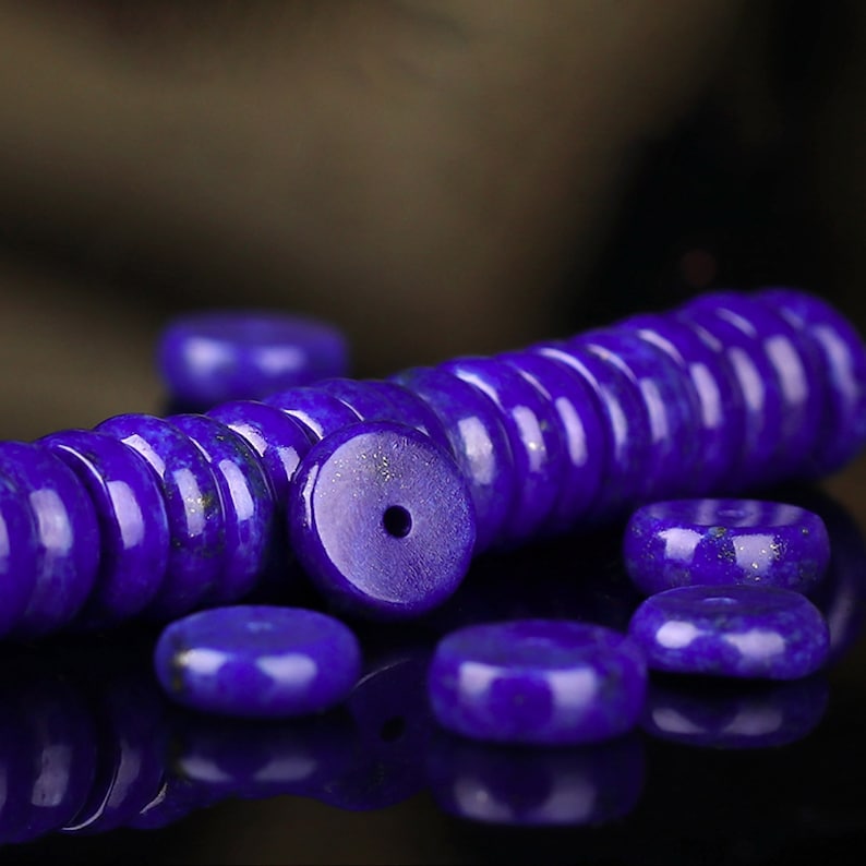 May include: A close-up of a pile of deep blue lapis lazuli beads. The beads are round and have a smooth, polished finish. They are arranged in a pile, with some beads scattered around the edges.